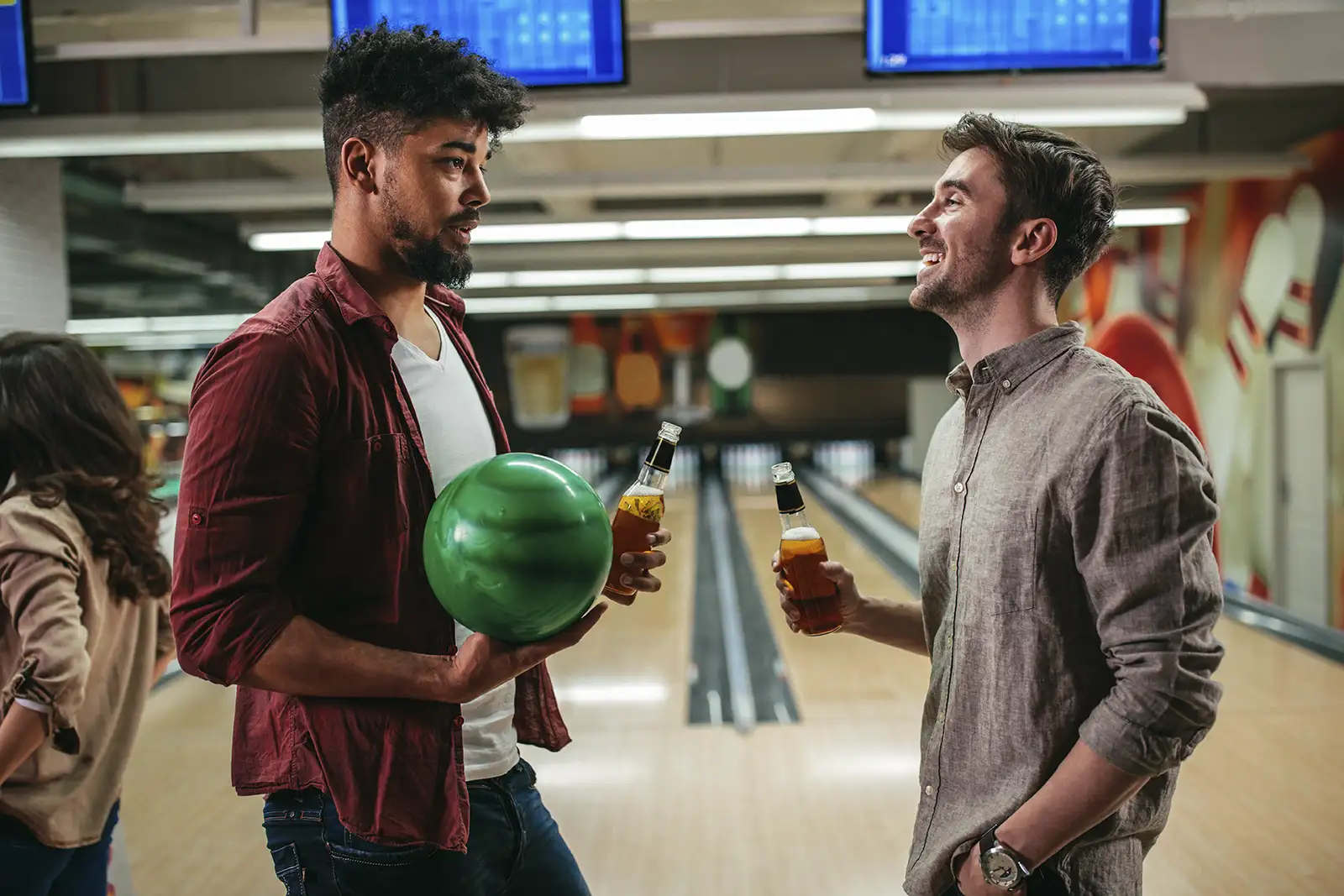 Two friends having a conversation at a bowling alley while holding beer bottles